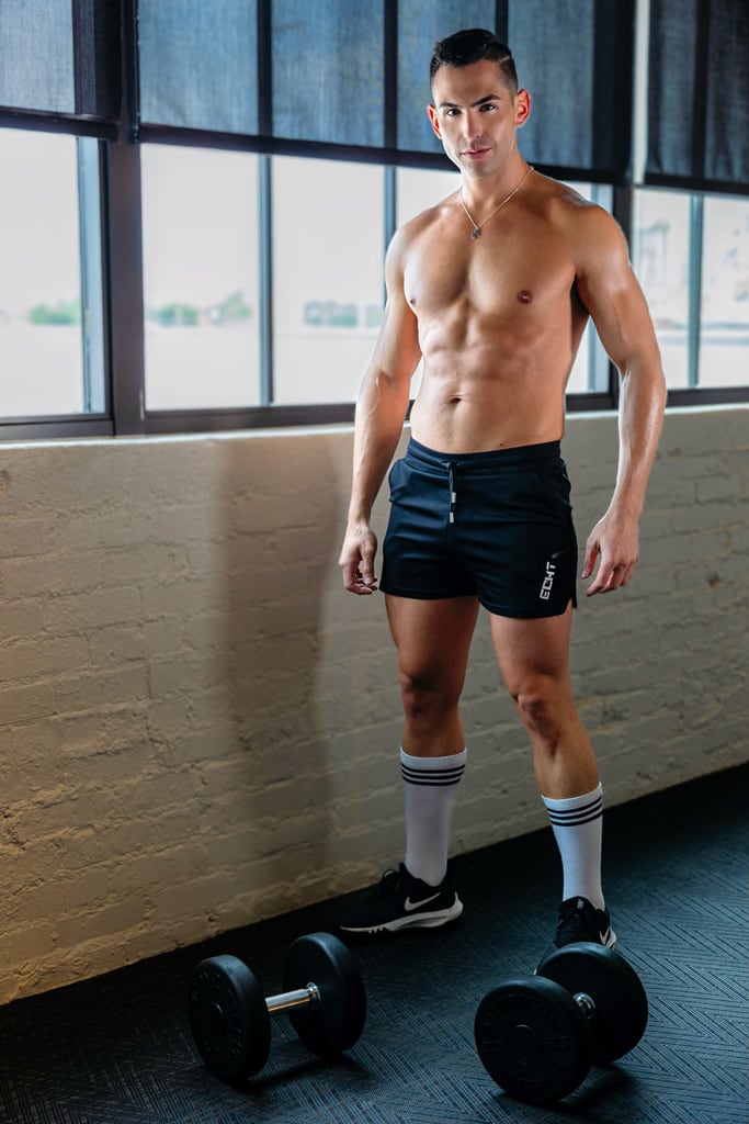 Coach is posing in front of windows topless and having dumbbells on the floor.